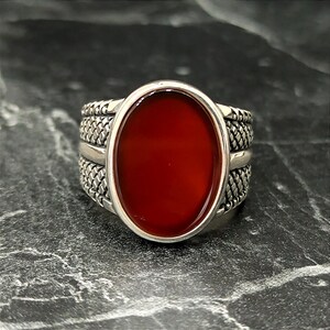 May include: A silver ring with a red carnelian stone set in an oval bezel. The band is decorated with a braided pattern.