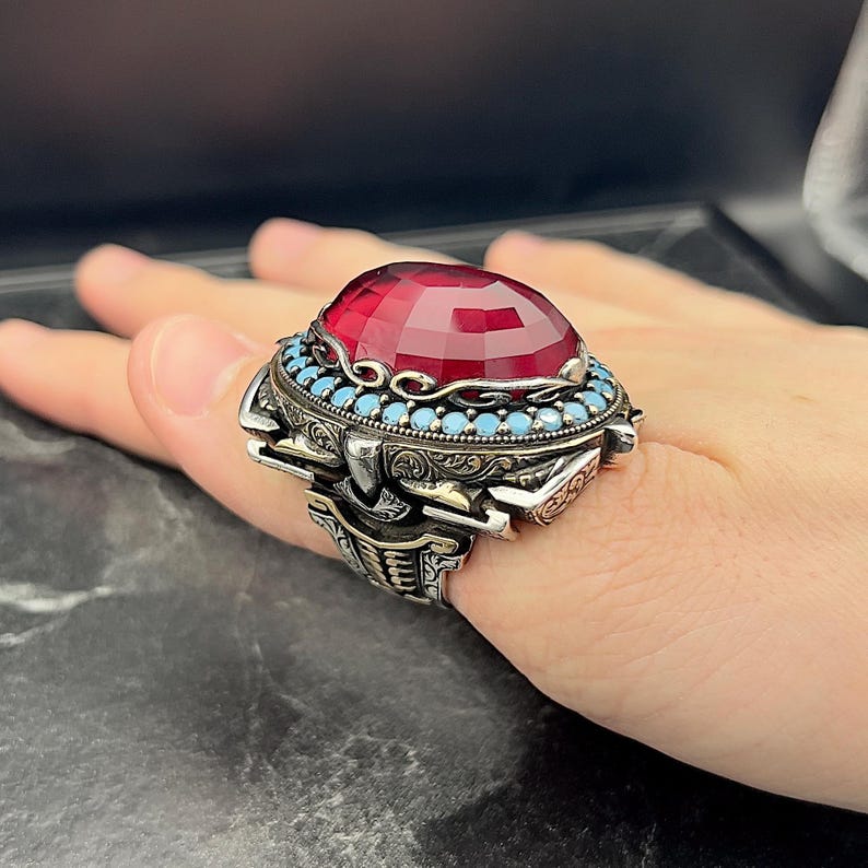 May include: A silver ring featuring a large, red gemstone encircled by a band of smaller, blue gemstones. The ring is adorned with elaborate, detailed engravings.
