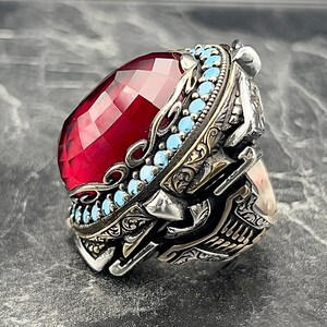 May include: A silver ring featuring a large, faceted, red gemstone encircled by a band of turquoise stones. The ring boasts intricate, ornate details and a skull-like design.