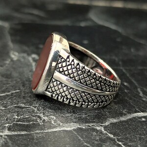 May include: A silver ring with a red oval gemstone set in a textured band. The band has a decorative design with a raised line.