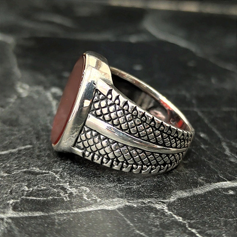 May include: A silver ring with a red oval gemstone set in a textured band. The band has a decorative design with a raised line.