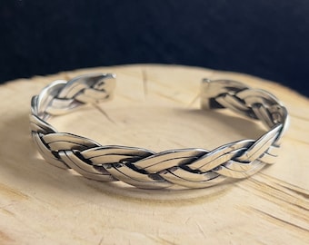 May include: A silver braided bangle bracelet with a woven design. The bracelet is open and has a flexible fit.
