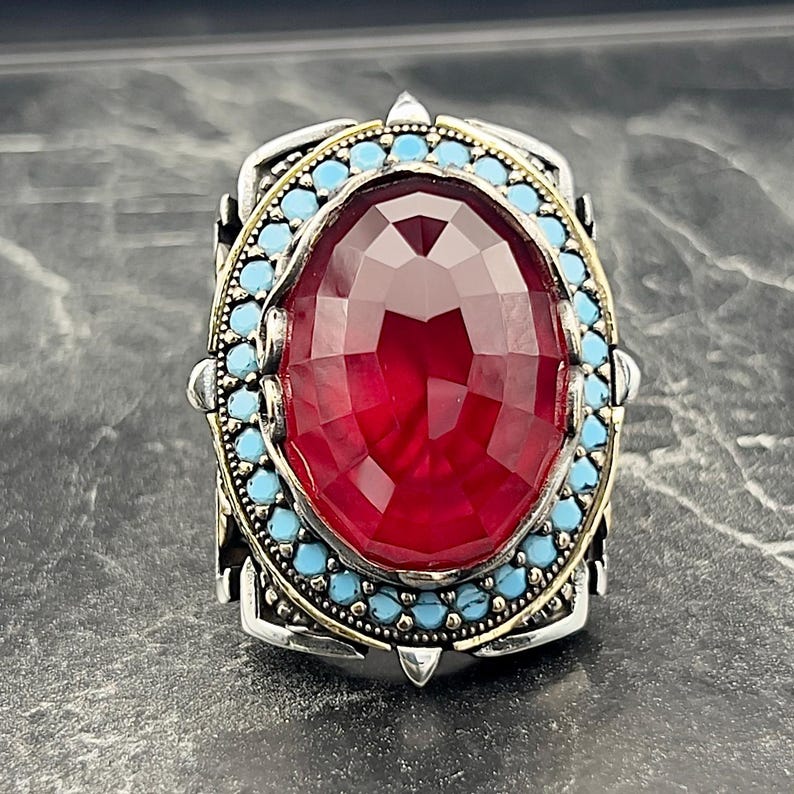 May include: A silver ring featuring a large red gemstone set within a border of turquoise stones.