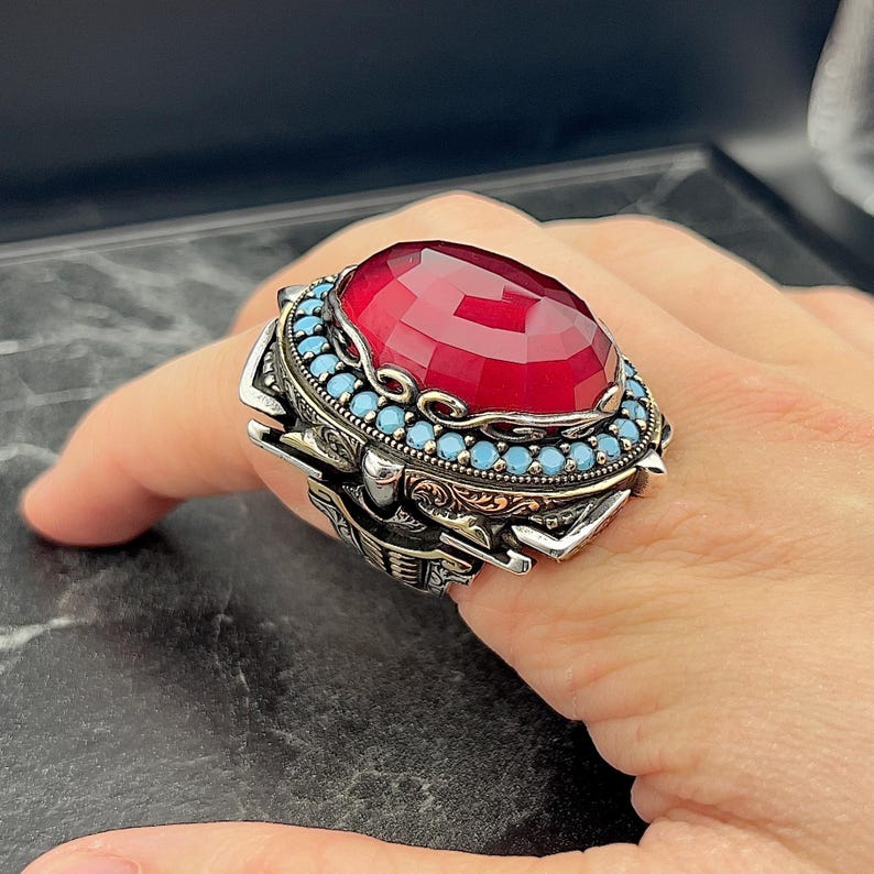 May include: A silver ring featuring a large red gemstone surrounded by a border of smaller turquoise gemstones. The ring has intricate details and a detailed design.