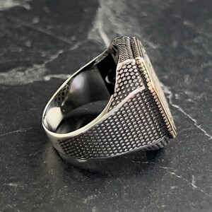 May include: A silver ring with a textured band and a square-shaped top. The top of the ring has a detailed design.