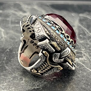 May include: A silver ring featuring intricate engravings and a large, red gemstone set in a gold-toned bezel. The ring is decorated with turquoise-coloured accents and the number "925" is visible on the band.