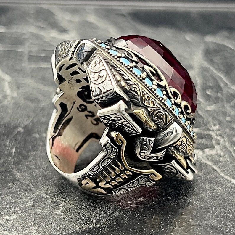 May include: A silver ring featuring intricate engravings and a large, red gemstone set in a gold-toned bezel. The ring is decorated with turquoise-coloured accents and the number "925" is visible on the band.