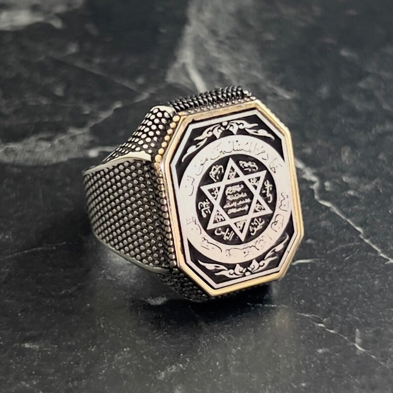 May include: A silver ring with a black octagonal center featuring a silver six-pointed star with Arabic script around it. The ring has a textured band.