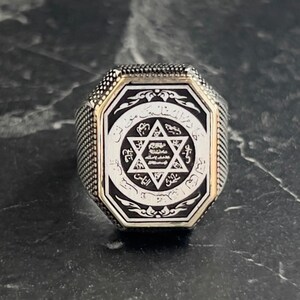 May include: A silver ring with an octagonal face featuring a black enamel inlay. The inlay depicts a six-pointed star with Arabic script surrounding it.
