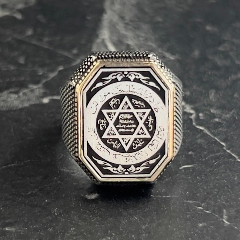 May include: A silver ring with an octagonal face featuring a black enamel inlay. The inlay depicts a six-pointed star with Arabic script surrounding it.