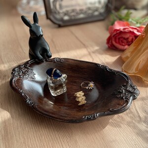 Könnte beinhalten: Ein dunkles braunes Holztablett mit einem dekorativen, verzierten Design. Eine schwarze Hasenfigur sitzt am Rand des Tabletts. Auf dem Tablett befinden sich drei goldene Ringe und ein kleines Parfümfläschchen.