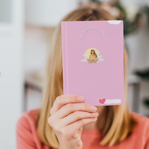 Może przedstawiać: Różowy dziennik medytacji z napisem "My Meditation Diary" i ilustracją osoby medytującej. Naklejka w kształcie serca znajduje się w prawym dolnym rogu. Dziennik jest trzymany przed osobą.