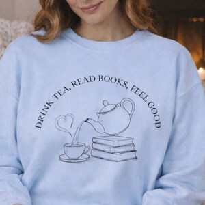 Könnte beinhalten: Hellblaues Sweatshirt mit dem Text "DRINK TEA, READ BOOKS, FEEL GOOD" und einer Teekannen- und Tassengrafik. Eine Person liest ein Buch, während sie auf einem Sofa sitzt. Eine Tasse und eine Kerze stehen auf einem Tisch.