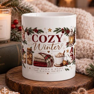 Könnte beinhalten: Ein weißer Keramikbecher mit einer festlichen Winterillustration und dem Text COZY Winter sowie SLOW DOWN & ENJOY THE LITTLE THINGS. Das Design zeigt eine Laterne, einen Stapel gefalteter Decken, eine Tasse heiße Schokolade und Tannenzweige.