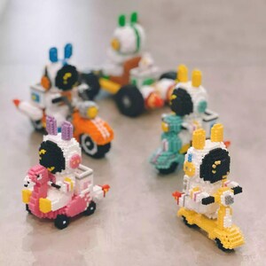 May include: Five colourful toy figures riding on scooters. Each figure is a white and black astronaut with a different coloured scooter. The scooters are pink, yellow, blue, orange, and green.