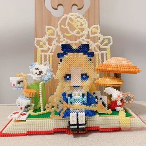 May include: A beaded figure of Alice from Alice in Wonderland sitting on a book with a blue dress, blonde hair, and a blue bow. The book is open and has a green field with a mushroom, a teacup, and a rabbit.