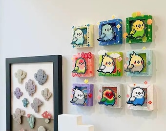 May include: Nine framed pixel art pieces featuring different colored birds. The frames are all different colors and have a white background. The frames are arranged in a grid pattern on a white wall.