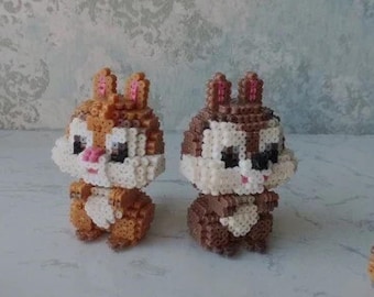 May include: Two small, brown and white, bead-crafted figurines of chipmunks. The chipmunks are standing on a white surface. A brown bead-crafted acorn is on the right side of the image.
