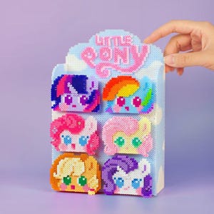 以下が含まれることがあります： 6つのカラフルなピクセル化されたリトルポニーのキャラクターの顔が特徴のディスプレイ。ディスプレイは水色で、ピンク色の「Little Pony」のロゴが付いています。各ポニーの顔は、ピンク、紫、黄色、青など、異なる色です。