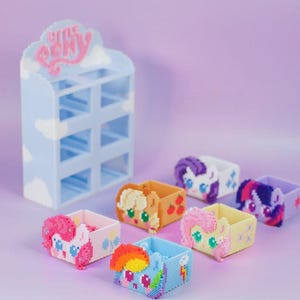 以下が含まれることがあります： ピンク色の「Little Pony」の文字が入った水色の収納棚。6つのカラフルなピクセル化された箱にはポニーのデザインが施され、棚の前に配置されています。箱はさまざまな色とポニーのキャラクターを特徴とし、柔らかな紫色の背景に設定されています。