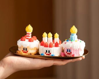 May include: Three miniature birthday cakes made of colorful beads. The cakes are decorated with candles and frosting. The cakes are on a gold tray.