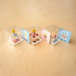 May include: Four miniature birthday cakes made of colorful plastic blocks in clear plastic boxes with a Happy Birthday card. Each cake has a different colored candle and frosting.