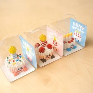 May include: Four miniature birthday cakes made of colorful plastic building blocks are displayed in clear plastic boxes with a handle. Each box has a card with the text "Happy Birthday" on it.
