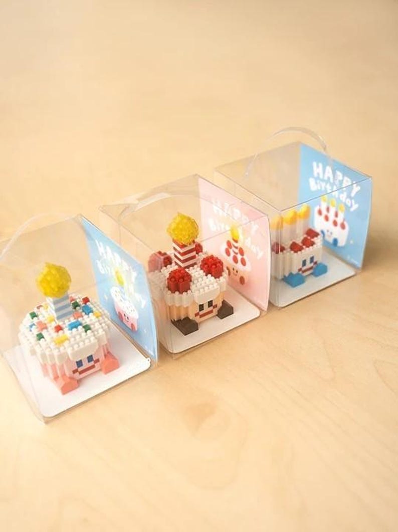 May include: Four miniature birthday cakes made of colorful plastic building blocks are displayed in clear plastic boxes with a handle. Each box has a card with the text "Happy Birthday" on it.