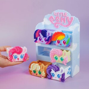 以下が含まれることがあります： マイリトルポニーのキャラクターが描かれた、カラフルなピクセル化された箱が6つ並んだディスプレイ棚。 棚は水色で雲のデザインで、「LITTLE PONY」の文字がピンク色で書かれています。 箱の一つは手で持たれています。