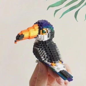 Pode incluir: Um tucano feito de contas coloridas. O tucano tem um corpo preto, um peito branco, um bico laranja e penas azuis e verdes na cabeça e na cauda.