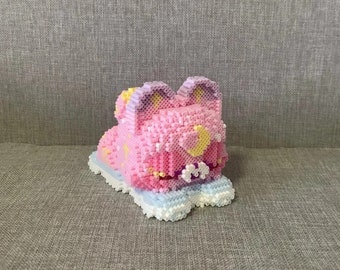 May include: A pink and purple beaded animal figurine with a yellow crescent moon and stars. The figurine is sitting on a white beaded base.