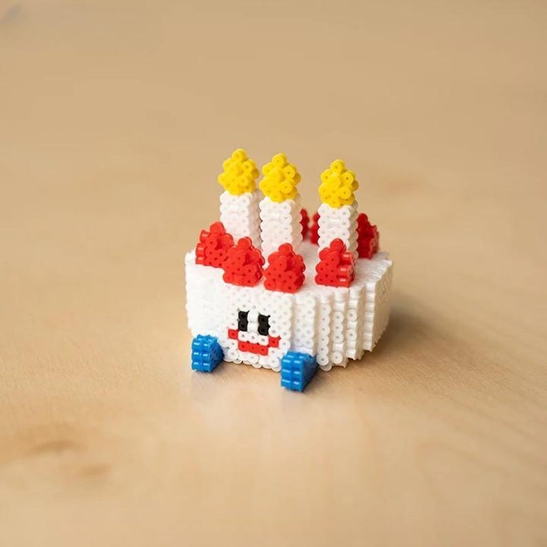 May include: A white, red, and yellow birthday cake made of perler beads. The cake has a smiling face and four candles on top.