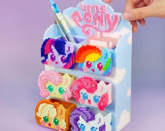 May include: A light blue desk organizer featuring six compartments, each adorned with a different My Little Pony character. The organizer has a cloud-shaped top with the words "Little Pony" in pink. A pen is placed in one of the compartments.