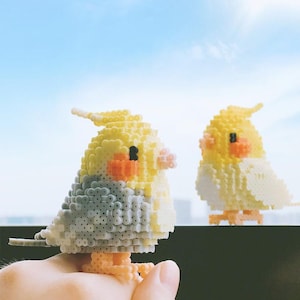 以下が含まれることがあります： 黄色と灰色のビーズでできた小さな鳥の置物2つ。左側の鳥は手で持たれている。右側の鳥は黒い表面の上に座っている。