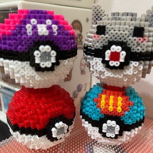 May include: Two Pokeball-themed pixel art figures. The first figure is a purple and pink Pokeball with a red Pokeball on top. The second figure is a gray and white Pokeball with a blue and yellow Pokeball on top.