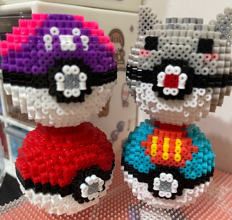 May include: Two Pokeball-themed pixel art figures. The first figure is a purple and pink Pokeball with a red Pokeball on top. The second figure is a gray and white Pokeball with a blue and yellow Pokeball on top.