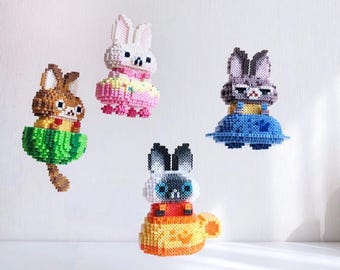 May include: Four colourful beaded bunny figurines with different expressions and outfits. Each bunny is sitting on a beaded platform with a different design. The platforms are in the colours green, blue, pink, and orange.