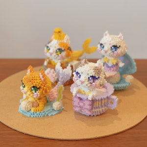 May include: Four adorable cat figurines made of colorful beads, each with a unique pose and expression. The cats are sitting on a light brown surface.
