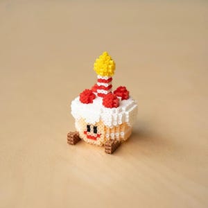 May include: A white, red, and yellow birthday cake made of beads with a smiling face and a lit candle on top.