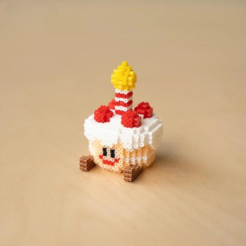 May include: A white, red, and yellow birthday cake made of beads with a smiling face and a lit candle on top.