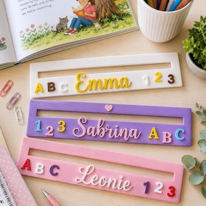 Könnte beinhalten: Drei rechteckige Namensschilder in Weiß, Lila und Rosa, jeweils mit einem horizontalen Schlitz, farbigen erhabenen Buchstaben für die Namen Emma, Sabrina oder Leonie sowie verschiedenen Zahlen. Die Schilder liegen neben einem Buch und Stiften.