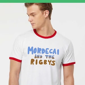 May include: A white ringer t-shirt with red trim on the collar and sleeve cuffs. The front of the shirt features the text "MORDECAI AND THE RIGBYS" printed in a stylized font, with "MORDECAI" in light blue and "AND THE RIGBYS" in brown lettering.