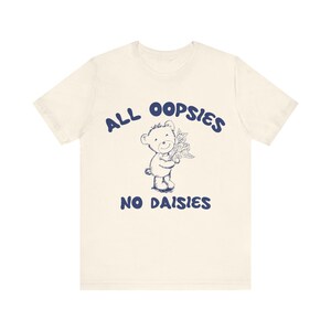 May include: A cream-colored t-shirt with a blue graphic of a teddy bear holding flowers. The text "ALL OOPSIES NO DAISIES" is printed in blue on the shirt.