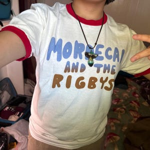 May include: A white t-shirt with red trim on the sleeves and collar. The shirt has a blue and brown graphic that says "Morecai and the Rigby's". A person is wearing the shirt and has a mushroom-shaped necklace on.