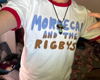Puede incluir: Una camiseta blanca con ribetes rojos en las mangas y el cuello. La camiseta tiene un gráfico azul y marrón que dice "Morecai and the Rigby's". Una persona lleva la camiseta y tiene un collar con forma de hongo.