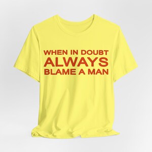 Może przedstawiać: Ż&oacute;łta koszulka z czerwonym napisem "WHEN IN DOUBT ALWAYS BLAME A MAN". Koszulka wykonana jest z miękkiego materiału i ma klasyczny okrągły dekolt. Tekst jest wyśrodkowany na przodzie koszulki.