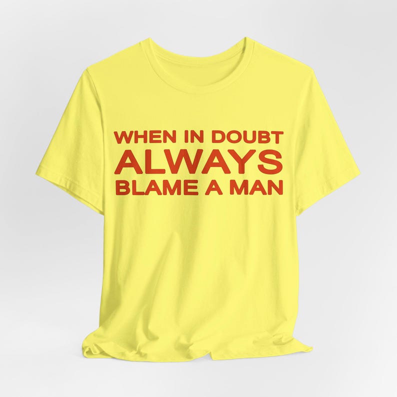 Może przedstawiać: Ż&oacute;łta koszulka z czerwonym napisem "WHEN IN DOUBT ALWAYS BLAME A MAN". Koszulka wykonana jest z miękkiego materiału i ma klasyczny okrągły dekolt. Tekst jest wyśrodkowany na przodzie koszulki.
