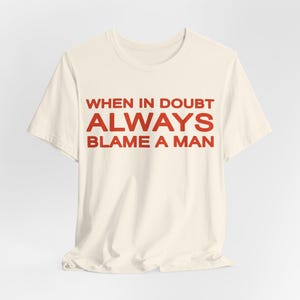 Może przedstawiać: Kremowy t-shirt z czerwonym napisem "WHEN IN DOUBT ALWAYS BLAME A MAN." Koszulka ma kr&oacute;tkie rękawy i okrągły dekolt. Tekst jest wyśrodkowany na przodzie koszulki.