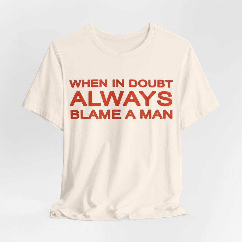 Może przedstawiać: Kremowy t-shirt z czerwonym napisem "WHEN IN DOUBT ALWAYS BLAME A MAN." Koszulka ma kr&oacute;tkie rękawy i okrągły dekolt. Tekst jest wyśrodkowany na przodzie koszulki.