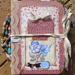 Pode incluir: Um diário feito à mão com um design floral. A capa apresenta um padrão floral rosa e branco, acabamento em renda e uma pintura em aquarela de flores azuis. Uma fita creme envolve o diário e um berloque de contas pende da lateral.